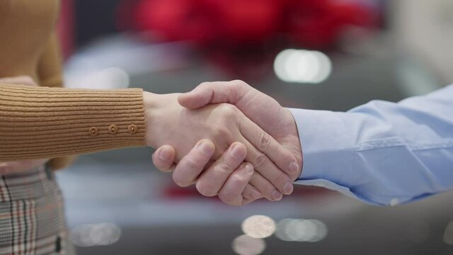 Close-up handshake of dealer and client in car showroom. Unrecognizable man and woman passing key in car dealership. Caucasian buyer and seller making deal. Slow motion.