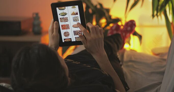 Young woman lying down in sofa at home and online orders fast food fom home. Online shopping from home. 