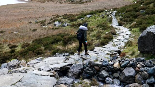 City woman with backpack walking carefully down the slippery stone path by lake Llyn Idwal in North Wales mountains.
