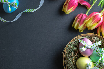 Easter symbol. Colorful egg with tape ribbon, spring tulips on dark rough stone background in Happy Easter decoration. Congratulatory easter flat lay design.
