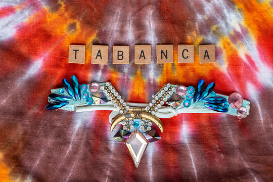 Tabanca, Spelled Out In Wooden Letters, With A Bedazzled Blue And Pink Diamond Costume Headpiece From Trinidad And Tobago Carnival. Multicoloured Tie Dye Fabric Background.