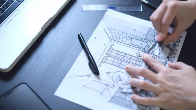The hand of an architect drawing a perspective of the bedroom with a black pen. On a black wooden desk where a tablet and pencil are placed.
