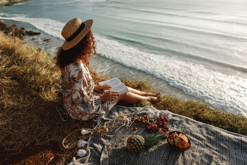Back view young curly hair brunette girl sit on sea cliff with open book in her hands, enjoy view and reading. Vacation by the sea