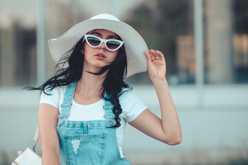 Beautiful brunette girl in  Cat Eye sunglasses and white  hat is looking into camera. She is outside. It is summer and hot weather and windy. Portrait. Soft colors preset.