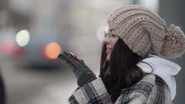 Portrait of a young dark-haired woman in round glasses and a knitted hat standing on the street on a winter day, talking on the phone, smiling, people are walking by.