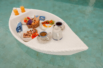Floating breakfast set in tray with fried egg, waffles bread , fresh fruits, mango juice  and coffee  in the swimming pool