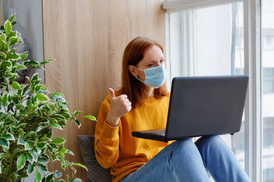 A female employee or student sits at the window with a mask on her face and a laptop. Cozy and convenient work remotely.