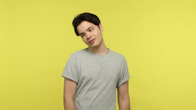 Crazy stupid young guy in casual gray t-shirt looking around with crossed eyes sticking out tongue, making silly brainless dumb face. Indoor studio shot isolated on yellow background