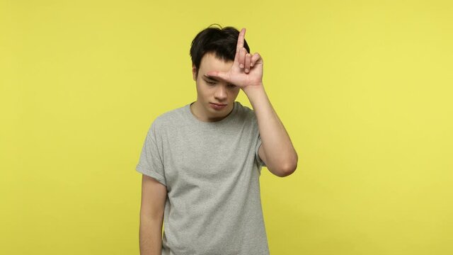 Frustrated unlucky young man in gray t-shirt standing showing loser gesture, upset about dismissal, lost job, failure in relations, life difficulties. Indoor studio shot isolated on yellow background