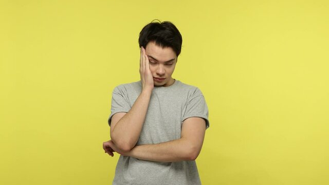 Frustrated bored teenager in casual gray t-shirt leaning head on hand listening boring talk, apathetic emotions, laziness and lack of energy. Indoor studio shot isolated on yellow background