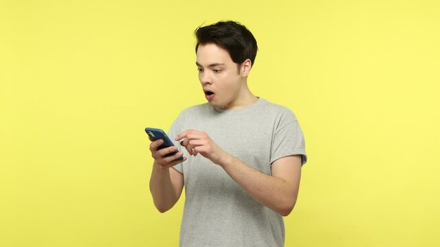 Surprised young man in casual gray t-shirt scrolling web pages on smartphone, looking with astonishment on discounts, shopping online. Indoor studio shot isolated on yellow background