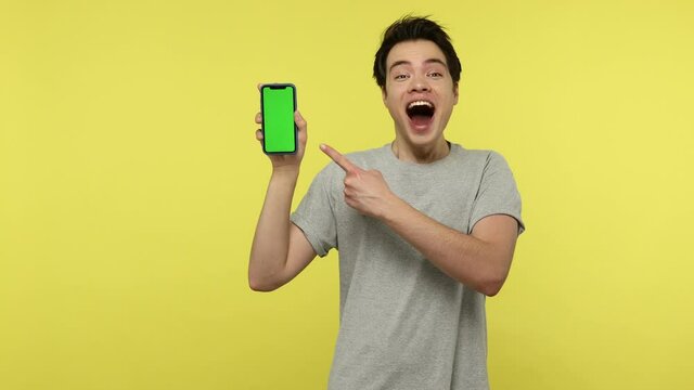 Extremely Happy Excited Teenager In Gray T-shirt Shocked With Cool Application On His Green Screen Smartphone, Looking At Camera Showing Thumbs Up. Indoor Studio Shot Isolated On Yellow Background