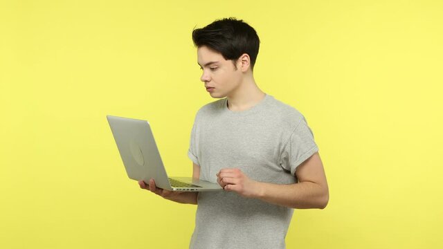 Concentrated young man student in casual gray t-shirt typing on laptop, working on his diploma project, writing article or programming. Indoor studio shot isolated on yellow background
