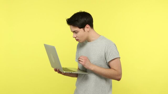 Shocked young man in gray t-shirt having problems working on laptop, disappointed with broken computer and data lost, deadline. Indoor studio shot isolated on yellow background