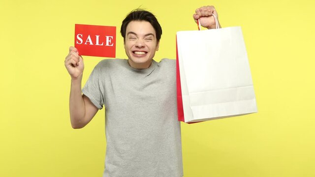 Happy excited young man in casual gray t-shirt bragging with shopping bags holding sale sign, extremely happy satisfied with low cost purchases. Indoor studio shot isolated on yellow background