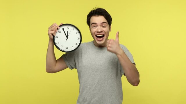 Look at time! Happy smiling young man in casual gray t-shirt holding big wall clock and looking at camera with satisfaction, finishing matters in time. Indoor studio shot isolated on yellow background