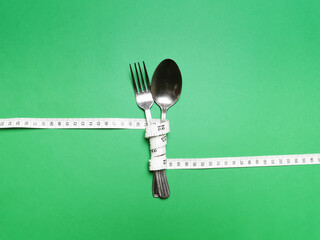 Spoon and knife are wrapped in white measuring tape on green background. Diet eating concept.Copy...