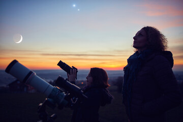 Mother and daughter observing stars, planets, Moon and night sky with astronomical telescope.