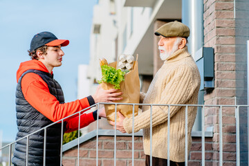 Young male courier in red uniforms bringing packet with fresh food to retired old man. Caucasian...