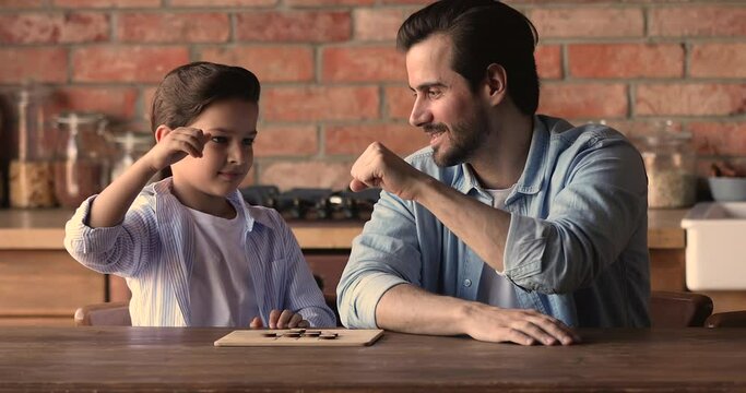 Happy small kid boy giving high five, bumping fists with smiling caring father, celebrating draw or win in checkers, playing game on wooden board, enjoying weekend family pastime together at home.