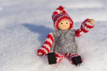 Close up of little doll with winter clothes and hat sitting in the snow in the winter sun