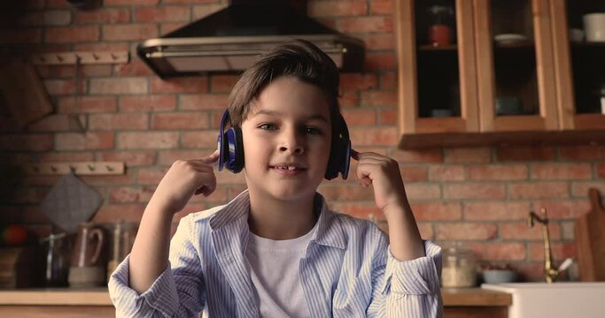 Adorable small 7s kid boy wearing wireless headphones, looking at camera, enjoying online class, holding video call meeting seminar training with teacher, distant education e-learning concept.
