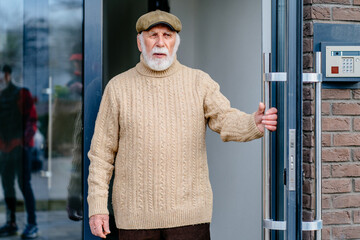 Old man in sweater and cap waiting someone on doorstep of his house. Lonely senior with gray beard...