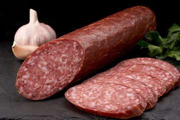 A stick of smoked sausage with slices of it and garlic. Macro shooting on a black background.