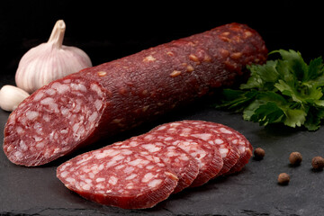 An appetizing stick of smoked sausage and slicing of it, on a slate board with a black background. Close-up.