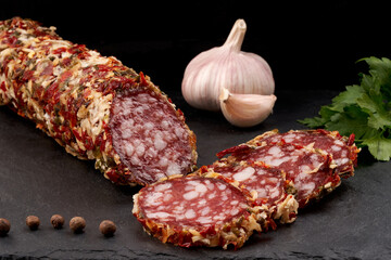 Appetizing smoked sausage stick in spicy sprinkle and sausage slicing on a slate board with a black background. Close-up.
