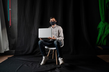 Man in a medical mask uses a laptop on a dark background