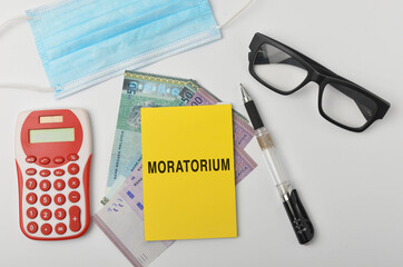 Top view of a banknotes, calculator, spectcles, face mask and pen with written MORATORIUM on white background. Selective focus.