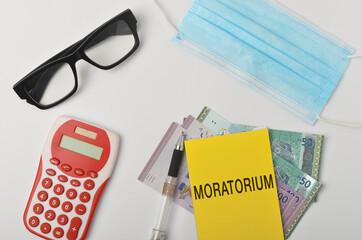 Top view of a banknotes, calculator, spectcles, face mask and pen with written MORATORIUM on white background. Selective focus.