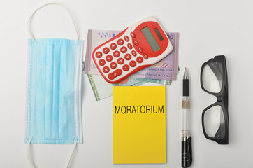 Top view of a banknotes, calculator, spectcles, face mask and pen with written MORATORIUM on white background. Selective focus.