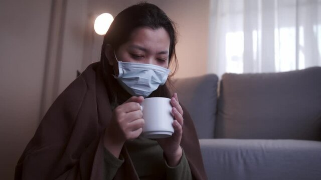 Young Asian woman drinking lukewarm water in times of suffering has chilling black eyelids all over her body due to a cold and uses masks and blankets to warm and quarantine herself