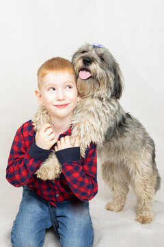 A Shaggy Mongrel Dog Embraces The Boy From Behind By The Neck. The Boy Holds His Pet By The Paws And Smiles Looking At The Camera