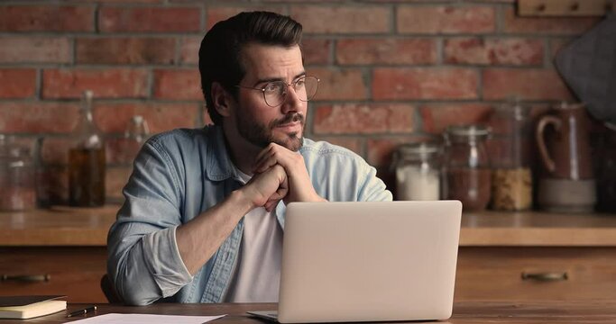 Pensive young businessman employee entrepreneur in eyeglasses looking in distance, thinking of problem solution or needs inspiration, typing working distantly on computer alone at home office.