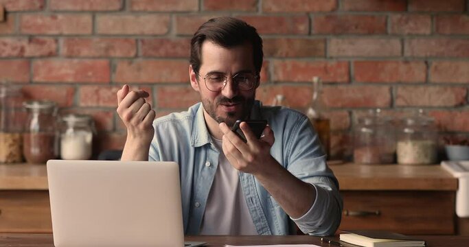 Happy young man in eyeglasses distracted from computer work holding loudspeaker mobile conversation, communicating distantly with colleagues, recording audio message or using voice assistant app.