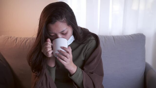 Young Asian woman drinking lukewarm water in times of suffering has chilling black eyelids all over her body due to a cold and uses masks and blankets to warm and quarantine herself
