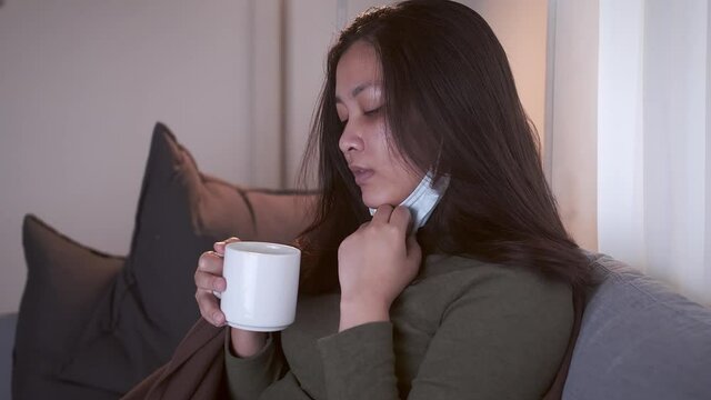 Sick Asian woman drinking lukewarm water in times of suffering has chilling black eyelids all over her body due to a cold and uses masks and blankets to warm and quarantine herself