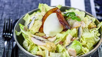 A serving of the famous Caesar Salad featuring, lettuce, croutons, anchovies, poached egg in a round bowl against a blue background
