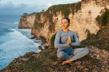 Beautiful woman doing yoga on a cliff, behind an amazing view in the ocean Bali Indonesia