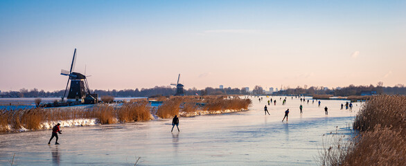 Classic Dutch scene with ice skaters on a lake during a cold period in winter. - obrazy, fototapety, plakaty