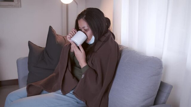 Young Asian woman drinking lukewarm water in times of suffering has chilling black eyelids all over her body due to a cold and uses masks and blankets to warm and quarantine herself