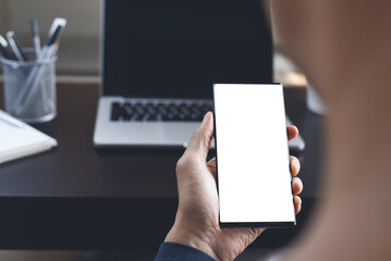 Mockup of man hand holding mobile phone with blank screen and working on laptop computer at office