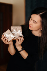 A beautiful brunette girl with long black hair in a black dress holds a golden gift box with a white ribbon bow