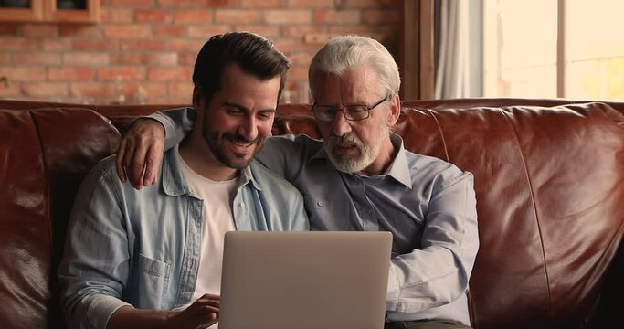 Happy bonding young 30s man and middle aged senior father looking at computer screen, reading email with good news, celebrating online lottery gambling win or watching football match, resting indoors.