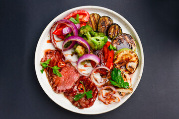 Grilled beefsteak and baked vegetables salad on a white plate over black background. Italian cuisine concept.