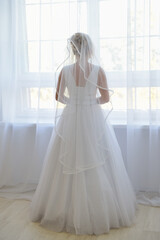 Naklejka premium Bride in a apartment in a wedding dress. Bride looks out of the window, wedding day. Charming bride stand in front of window in a white wedding dress.