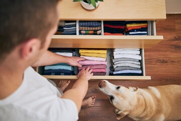 Organizing and cleaning home. Man preparing orderly folded t-shirts in drawer with his curious dog.